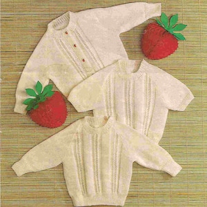 Puede incluir: Se muestran tres suéteres de punto color crema con detalles acanalados y cuellos redondos. Un suéter tiene pequeños botones rojos. También hay dos decoraciones de fresas rojas de felpa con hojas verdes. Los suéteres están colocados sobre una estera de bambú.