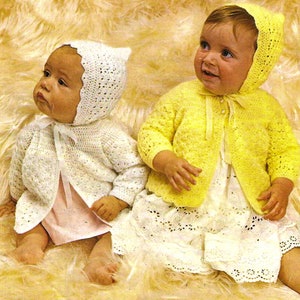 May include: Two babies wearing crocheted jackets and bonnets. One baby is wearing a white jacket and bonnet, while the other is wearing a yellow jacket and bonnet. Both babies are sitting on a fluffy white surface.