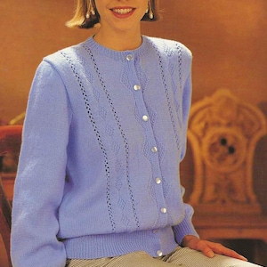 May include: A light blue cardigan with a round neckline and long sleeves. The cardigan features decorative openwork details and is fastened with silver buttons. The person is wearing patterned pants.