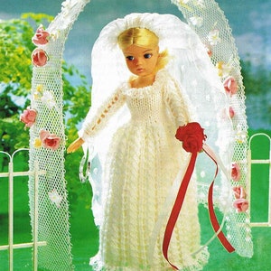 Puede incluir: Un vestido de muñeca blanco de punto con un borde de encaje y un velo. La muñeca sostiene un ramo de rosas rojas con una cinta blanca.