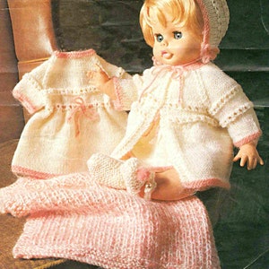 May include: A knitted doll outfit set in white and pink. The set includes a dress, cardigan, booties, and a hat.