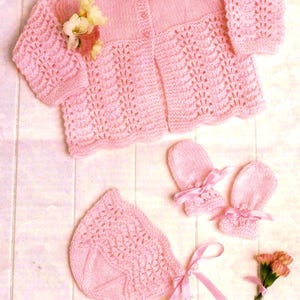 以下が含まれることがあります： ベビー用のピンク色のニットセット。カーディガン、帽子、ミトンが含まれています。カーディガンはボタン留めで、装飾的な透かし編みがあります。帽子とミトンはピンクのリボンで飾られています。小さな花のブーケが繊細さを添えています。