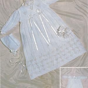 May include: A white knitted baby christening gown with matching bonnet and booties. The gown features long sleeves, a V-neck, and decorative ribbon accents. The bottom hem has a diamond pattern. The bonnet has a ribbon tie.