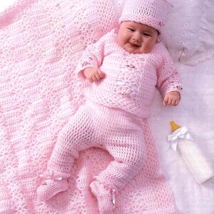 Puede incluir: Un conjunto de bebé de crochet rosa con un gorro a juego. El conjunto incluye una camiseta de manga larga, pantalones y un gorro. El conjunto está colocado sobre una manta de crochet rosa.