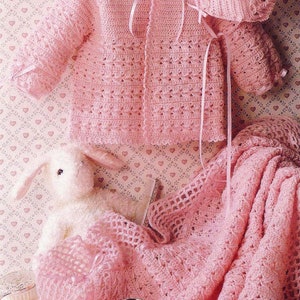 Puede incluir: Un conjunto de bebé de ganchillo rosa que incluye un cárdigan, un gorro, botines y una manta. El conjunto está colocado sobre una superficie con estampado rosa y blanco.