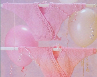 May include: Two pink knitted baby sweaters with a wrap style and a cable knit design. The sweaters are hanging on a white hanger against a light pink background.