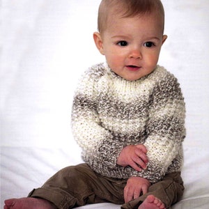 May include: A baby wearing a hand-knitted sweater in shades of white, brown, and gray, paired with brown pants. The baby is sitting on a white surface.
