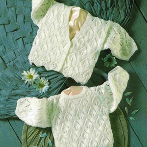 May include: Two white crocheted baby sweaters with a delicate lace pattern. The sweaters are laid out on a green woven surface.