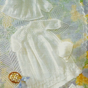 May include: A white knitted baby gown, jacket, booties, and hat set. The set is laid out on a white crocheted blanket with a floral pattern.