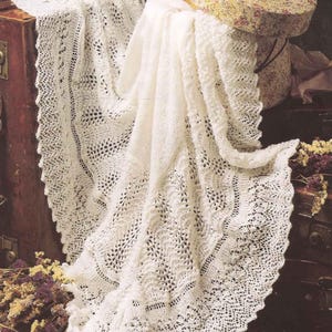 Peut inclure: Une couverture de bébé en crochet blanc avec un motif de dentelle délicat. La couverture est drapée sur une valise vintage et une boîte à motifs floraux.
