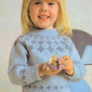 May include: A light blue knitted sweater with a high collar and diamond pattern. The sweater is worn by a young person holding a small bouquet of felt flowers. The flowers are in shades of pink, yellow, and purple.