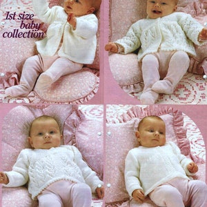 Puede incluir: Un cárdigan blanco de punto para bebé con un patrón de punto de cable. El cárdigan se muestra en un bebé que lleva pantalones rosas y está sentado en una almohada rosa con estampado floral blanco.