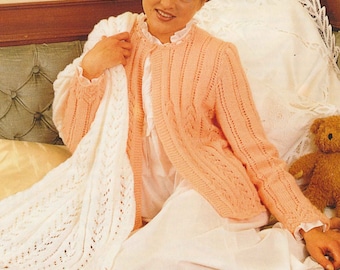 May include: A woman wearing a peach-colored knitted cardigan with a white lace trim. She is sitting on a bed with a white blanket and a teddy bear.