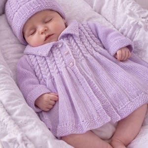 May include: A light purple knitted baby outfit consisting of a long-sleeved coat with a collared neckline, three front buttons, and vertical cable knit detailing, paired with a matching knitted beanie hat with a textured cable knit pattern.