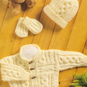 Puede incluir: Un conjunto de punto para bebé en color crema, que incluye un cárdigan con botones, un gorro con pompón y un par de manoplas.