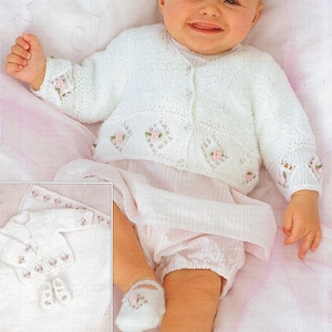 Può includere: Un cardigan bianco lavorato a maglia per neonato con un delicato motivo a pizzo e decorazioni floreali rosa. Il cardigan è abbottonato e ha maniche lunghe. Il bambino indossa un vestito bianco e scarpine bianche.