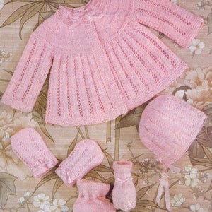 Puede incluir: Un conjunto de bebé de punto rosa que incluye un cárdigan, un gorro y botines. El cárdigan tiene un patrón de encaje y un lazo en el escote. El gorro y los botines también están hechos con un patrón de encaje.
