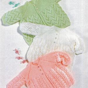 Può includere: Tre cardigan in maglia per neonati in diversi colori: verde, bianco e rosa. I cardigan sono disposti su una superficie bianca. DK o 4 fili, 14 - 20 pollici, 36 - 51 cm.