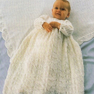 May include: A baby wearing a white knitted christening gown and matching blanket. The gown features a detailed, openwork pattern and long sleeves with ribbon ties. The blanket has a scalloped edge.