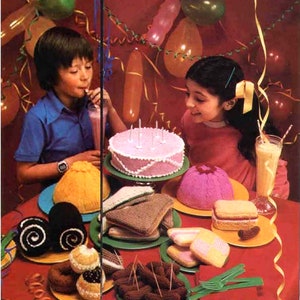 May include: A birthday party table setting with a pink cake, knitted food items, and a pink and yellow balloon backdrop.
