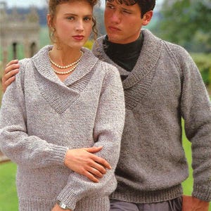 May include: A couple wearing matching gray knit sweaters with shawl collars. The woman wears a pearl necklace and a watch. The man has his arm around her. The sweaters have a textured appearance.