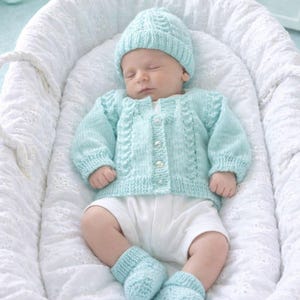 Könnte beinhalten: Ein helles mintgrünes Strickset für Babys, bestehend aus einer langärmeligen Strickjacke mit Knopfleiste und Zopfmuster, einer passenden Mütze und einem Paar Babyschuhen. Das Set wird über einem schlichten weißen Kurzarmbody getragen.
