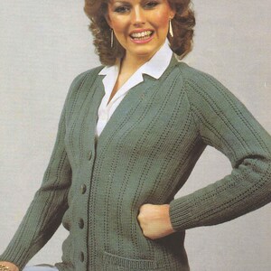 Pode incluir: Um cardigan verde de malha com botões e bolsos. O cardigan é usado sobre uma camisa branca com gola. A mulher na imagem está sorrindo e usando brincos e uma pulseira dourados.