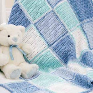 Op de afbeelding: Een witte teddybeer met een lichtblauw geruit lint om de nek en bijpassende stof op de pootjes, zittend naast een gehaakte deken met een vierkant ruitpatroon in tinten wit, lichtblauw en turquoise.