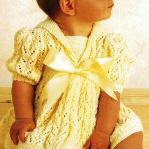 Pode incluir: Um bebé com um vestido de malha amarelo pálido com laço e meias a condizer. O vestido apresenta um padrão texturizado e mangas curtas. O bebé está sentado numa superfície de madeira.