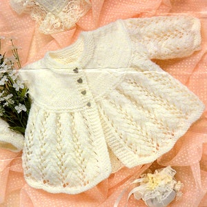 May include: A white knitted baby cardigan with a delicate lace pattern and button fastening. The cardigan has a scalloped hem and is laid on a pink background.