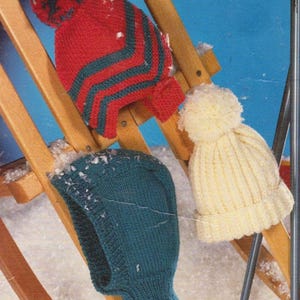 May include: Three knitted winter hats in a snowy scene. A red hat with green stripes and a pom-pom, a teal balaclava, and a cream-colored hat with a pom-pom are displayed on a wooden sled.