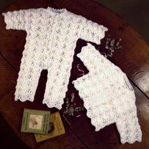 May include: A white, open-weave baby romper and matching jacket set. The garments are made of a delicate, lacy knit fabric with a scalloped edge. The set is displayed on a dark wooden surface with small books and decorative elements.