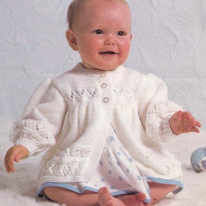 May include: A white knitted baby cardigan with a button closure and a delicate lace pattern. The cardigan has a ruffled hem and is worn by a baby sitting on a white surface.
