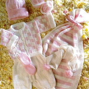 May include: A collection of baby knitwear in soft pink and white. The set includes a hat, booties, a romper, mittens, and a blanket. The blanket is striped, and the romper has a checkered pattern. The items are arranged on a bed of yellow and white flower petals.