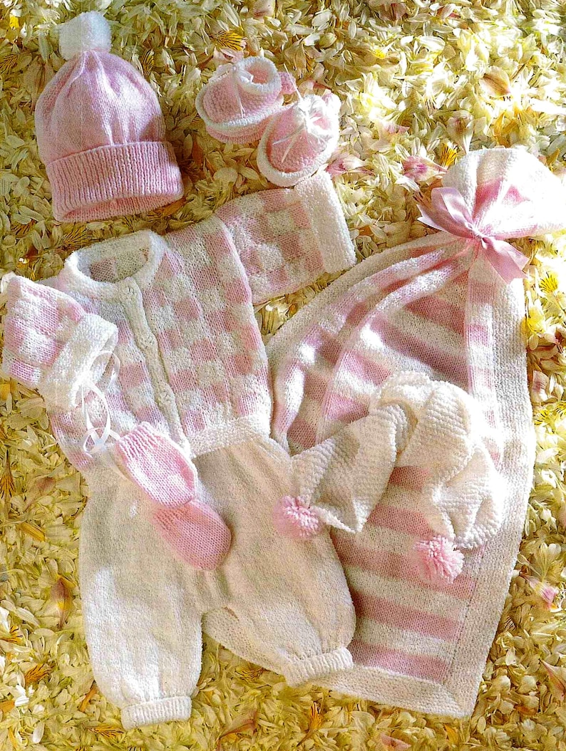 K&ouml;nnte beinhalten: Ein Set aus Baby-Strickwaren in Rosa und Wei&szlig;. Es beinhaltet eine M&uuml;tze, Babyschuhe, einen Strampler, F&auml;ustlinge und eine gestreifte Decke. Der Strampler hat ein Karomuster. Die Artikel liegen auf einem Bett aus gelben und wei&szlig;en Bl&uuml;tenbl&auml;ttern.