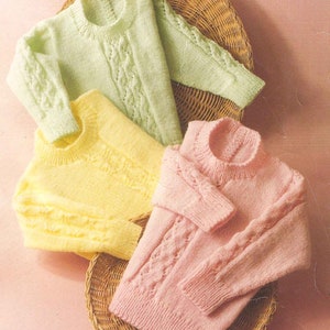 May include: Four knitted sweaters in pastel colors: light green, yellow, and pink. Each sweater has a crew neck and cable knit detailing on the sleeves and front. The sweaters are arranged on a woven basket, with a pink background.