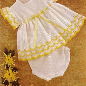 May include: A white knitted baby dress with yellow trim and a matching nappy cover. The dress has a ruffled neckline and a gathered skirt. The nappy cover has a simple design.