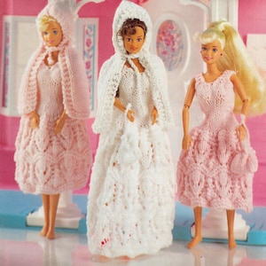 May include: Three dolls wearing knitted outfits. One doll is in a white hooded cape and dress, another in a pink hooded cape and dress, and the third in a pink dress. The outfits have a textured, openwork design.