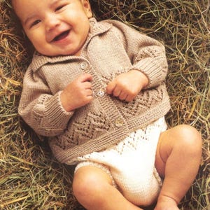 May include: A smiling baby wearing a light brown knitted cardigan with a decorative pattern and button closure. The baby is also wearing white knitted diaper covers. The baby is lying on a bed of hay.