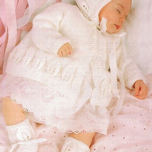 May include: A white knitted baby outfit with a matching hat and booties. The outfit features a delicate lace pattern and a satin ribbon.