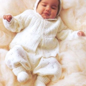 Puede incluir: Un bebé con un conjunto blanco de punto, que incluye una chaqueta con capucha, pantalones, gorro y calcetines. El conjunto tiene un patrón texturizado y está abotonado en la parte delantera. El bebé está acostado sobre una superficie suave y de color claro.