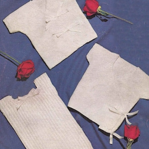 May include: Three white knitted baby sweaters with different styles and necklines. The sweaters are laid out on a blue background with red roses.