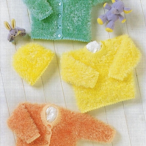 Puede incluir: Un surtido de suéteres y un gorro de bebé de punto esponjoso en tonos verdes, amarillos y naranjas. El suéter verde es una rebeca con botones. También se muestra un suéter y un gorro amarillos. Un peluche está al lado del suéter verde.
