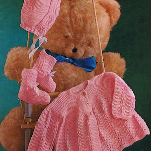May include: A pink knitted baby cardigan with a button fastening, a matching pink knitted hat with a bow, and pink knitted booties. The set is displayed on a teddy bear.