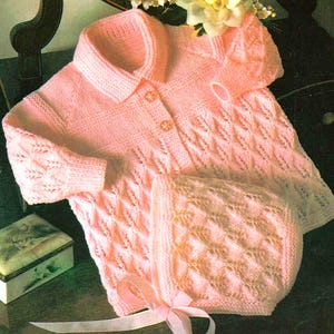 May include: A light pink knitted baby set, including a collared sweater with three buttons and a matching bonnet. The set features a diamond pattern and is tied with a pink ribbon. The set is made of soft yarn.