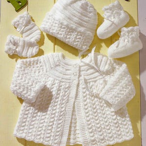 Puede incluir: Un conjunto de ropa de bebé blanco tejido a mano, que incluye una chaqueta, un gorro y patucos. El conjunto presenta un patrón texturizado y delicados detalles de cinta. La chaqueta tiene dos pequeños botones. Los patucos y el gorro tienen un borde festoneado.