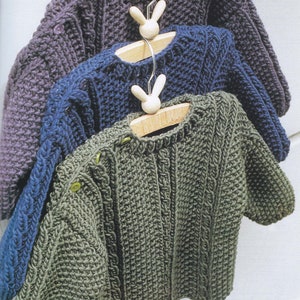 Puede incluir: Tres jerséis de punto para bebé en diferentes colores: morado, azul y verde. Los jerséis están colgados en perchas con ganchos en forma de conejo.