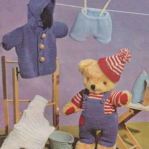 May include: A knitted teddy bear wearing blue dungarees, a red and white striped shirt, and a red and white striped hat. The bear is standing on a wooden stool next to a blue basket and a wooden ironing board with an iron on it. There are also knitted clothes hanging on a wooden clothes rack.