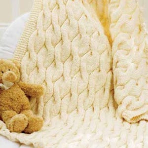 May include: Cream-colored knitted baby blanket with a cable knit design. A small, brown teddy bear with a cream-colored bow is resting on the blanket. The blanket is folded over a white surface.