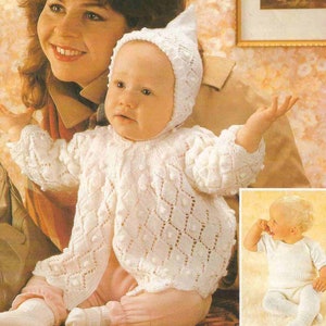 May include: A white knitted baby cardigan with a bobble stitch pattern and a matching bonnet. The cardigan has a button closure and a scalloped edge. The baby is wearing a pink onesie underneath.